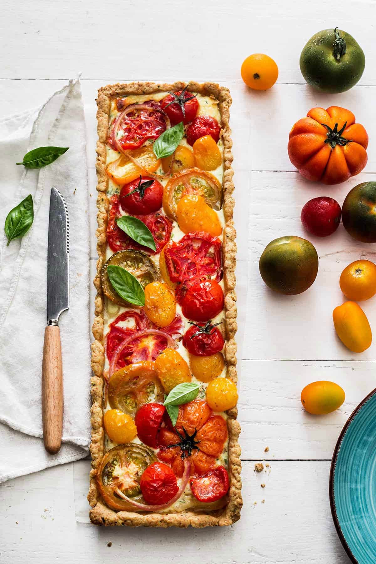 Crostata di pomodori, also known as Italian tomato pie on a rustic white table with heirloom tomatoes scattered around.