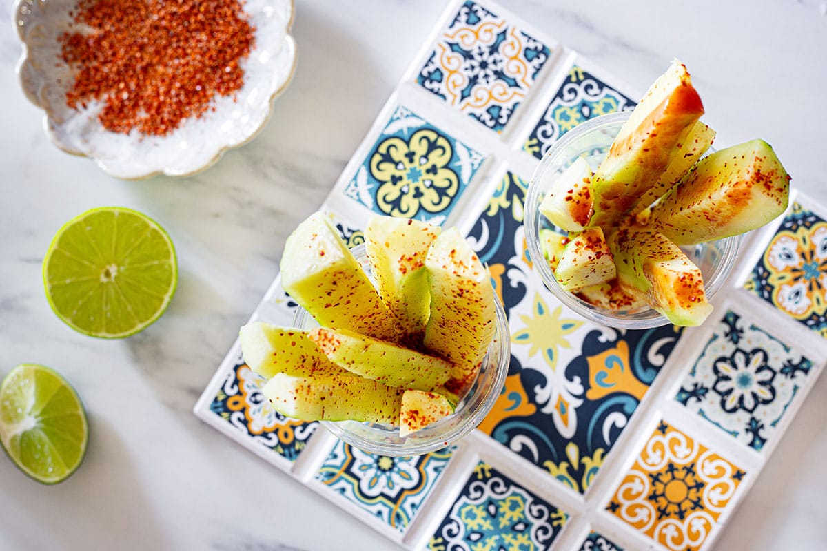 Two cups with cucumbers sticks sprinkled with chili powder and drizzled with hot salsa. A small plate with chili powder and lime wedges on the side.