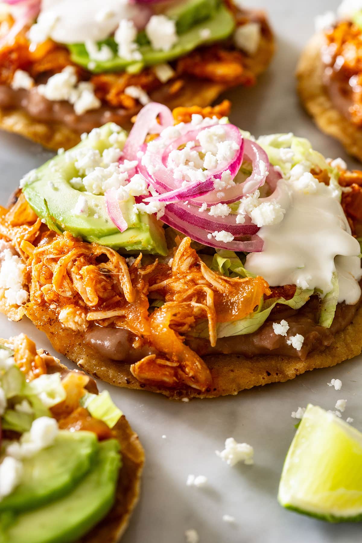 Close-up of a tinga tostada topped with lettuce, avocado, pickled onions, cream, and cheese.
