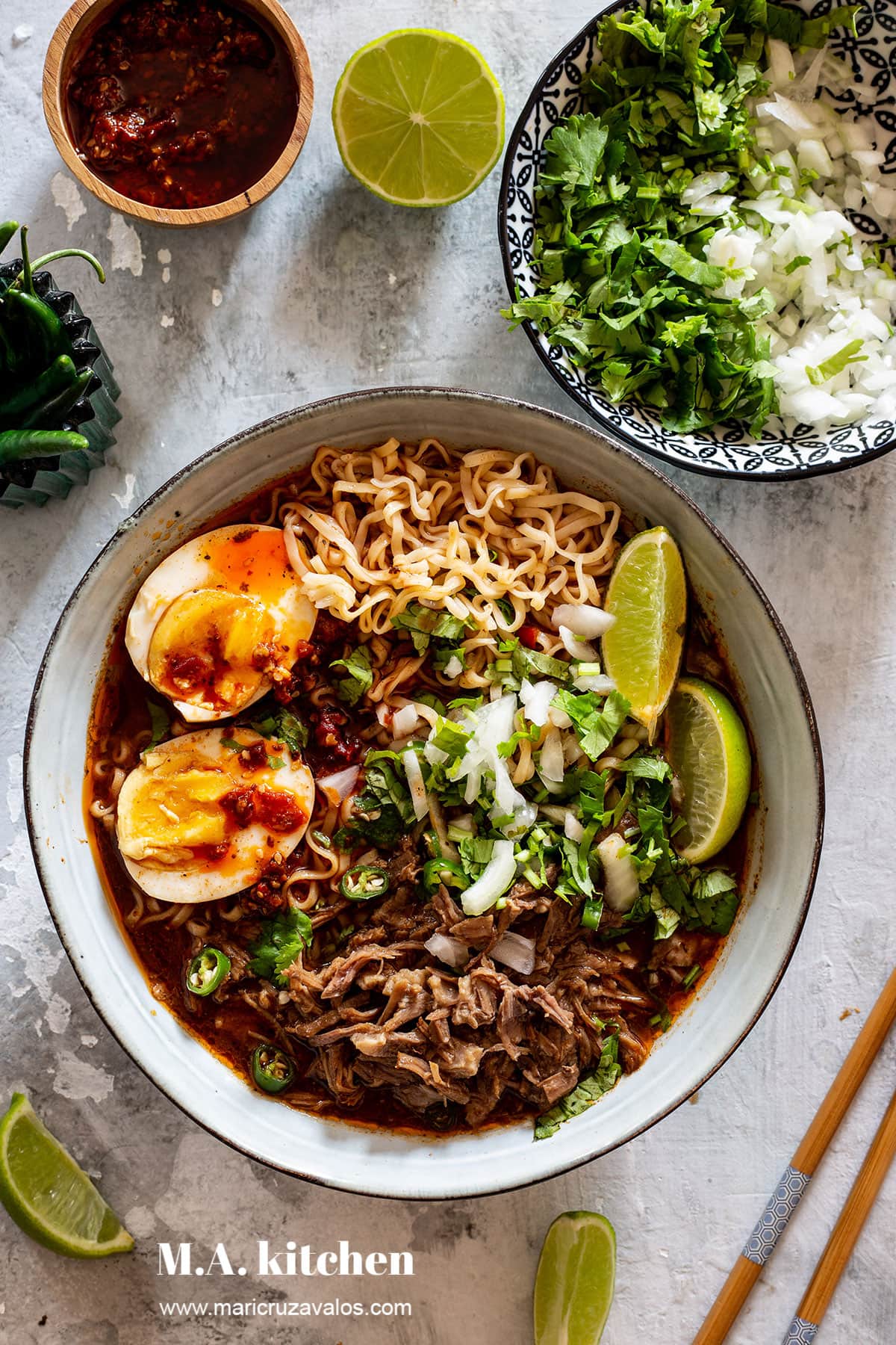 Birria ramen served with boiled eggs, cilantro, onion, chili oil and wedges of lime.