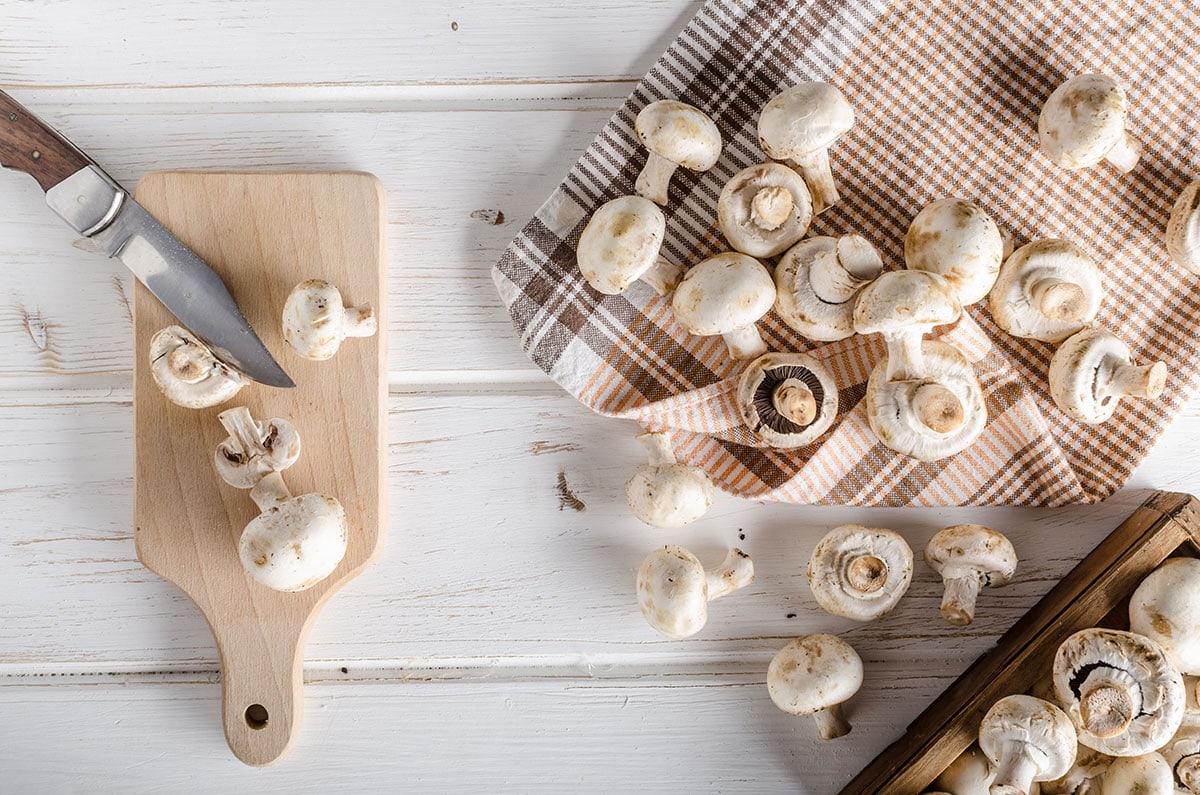 Champignon mushrooms on a white table scattered, and some on a cutting table with a knife near by.