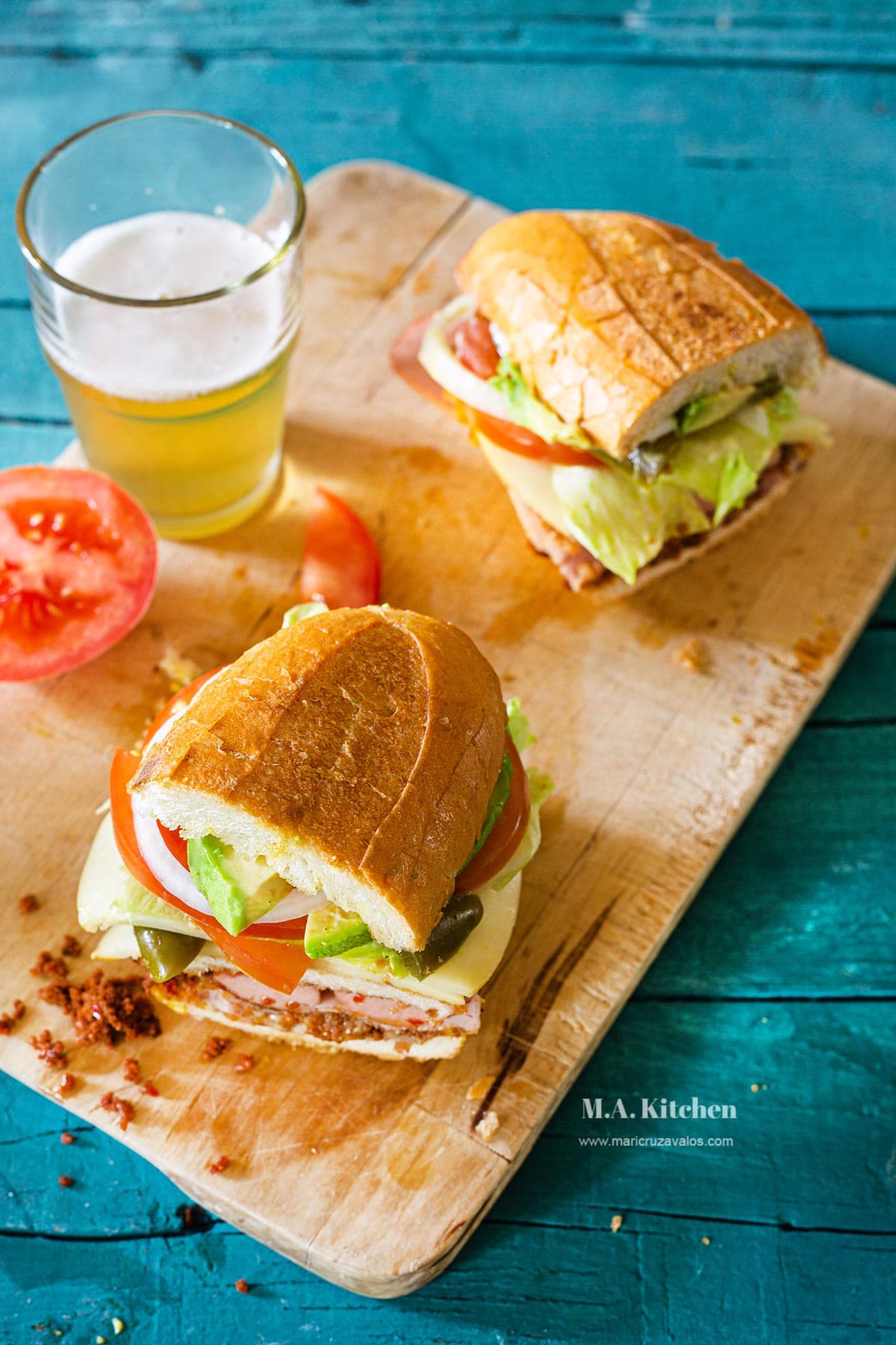 Mexican torta cubana cut in half in a cutting board with a glass of beer on the side.