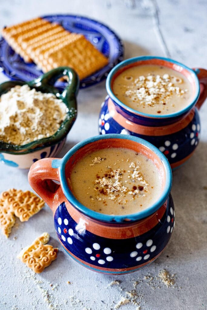 Atole de pinole served in Mexican ceramic mugs with cookies and pinole powder on the side.