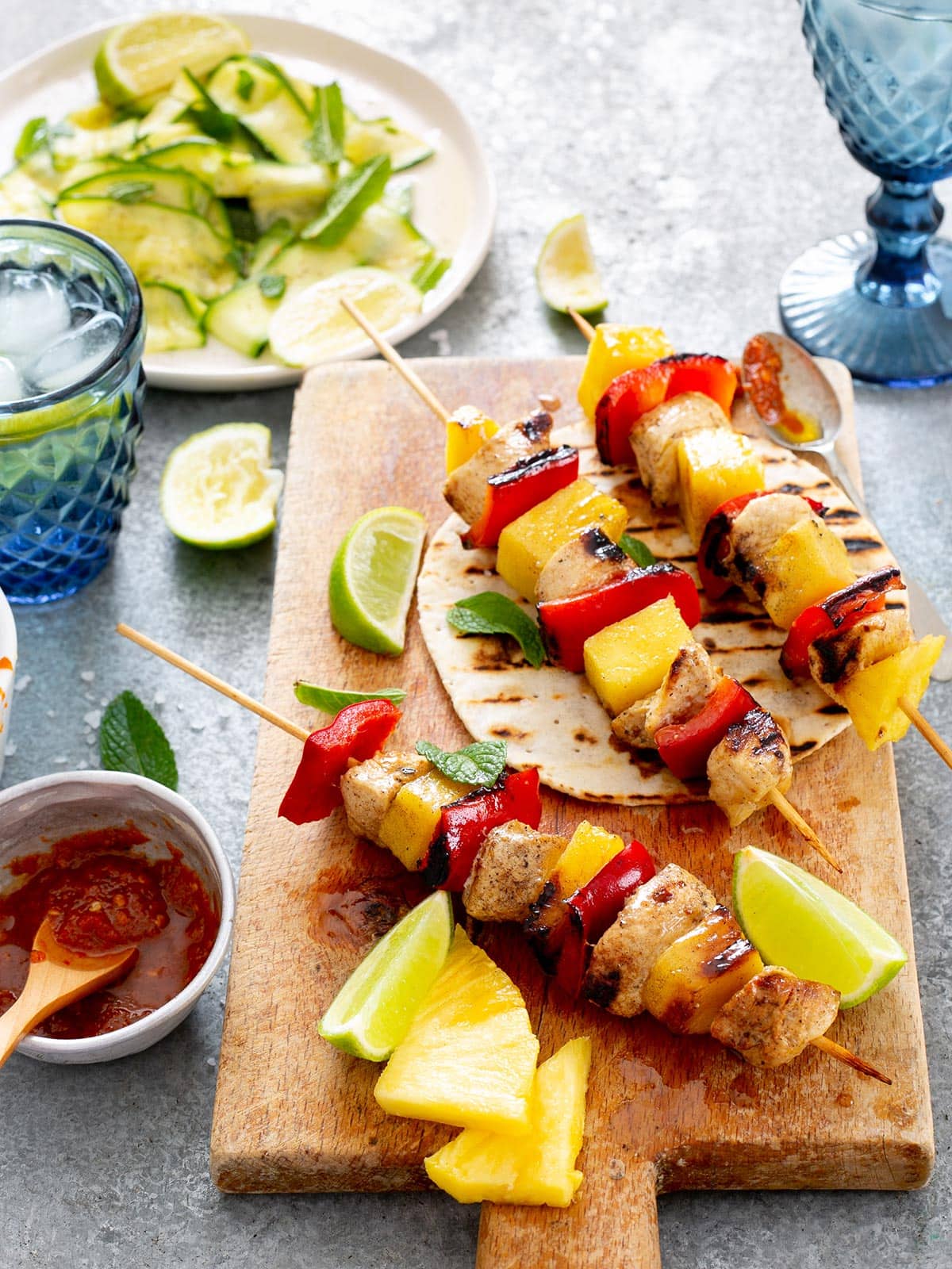 Brochetas de pollo on a wooden board with tortillas, lime wedges, salsa, and salad on the side.