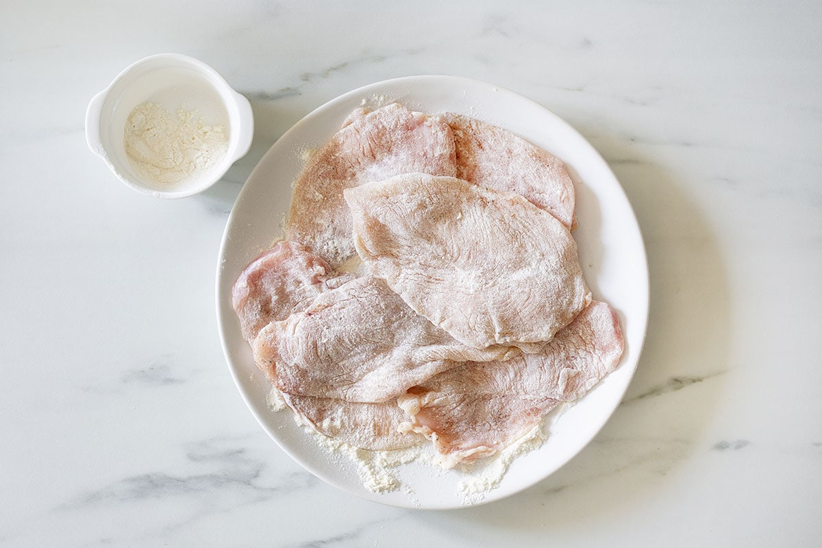 Chicken breast slices seasoned and dredged in flour and placed on a plate.