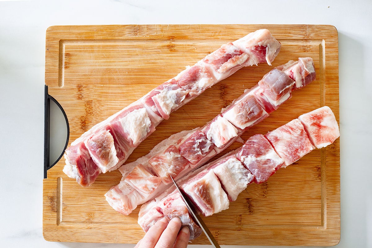 Pork belly cut into large strips on a cutting board and a hand showing how to make deep cuts on them.