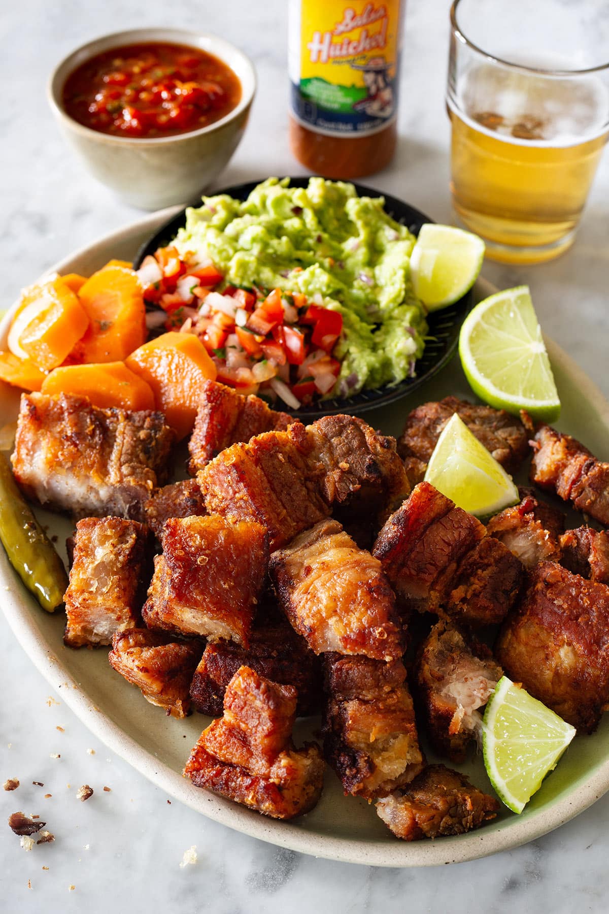 Mexican chicharrones in a platter with lime wedges, guacamole, pico de gallo, and pickled carrots. Behind a bowl with salsa, a bottle of hot sauce, and beer in a glass.