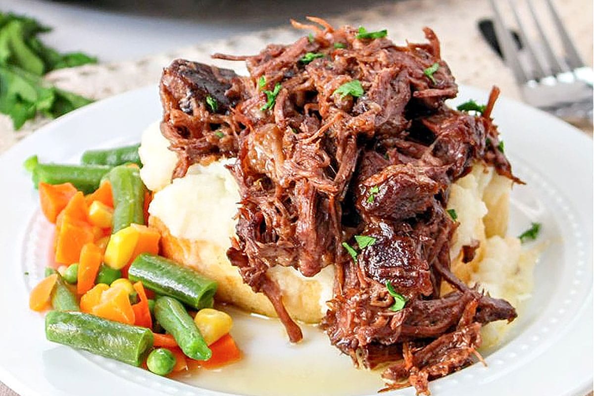 Slow cooker beef manhattan served on top of creamy mashed potatoes with mixed veggies on the side.
