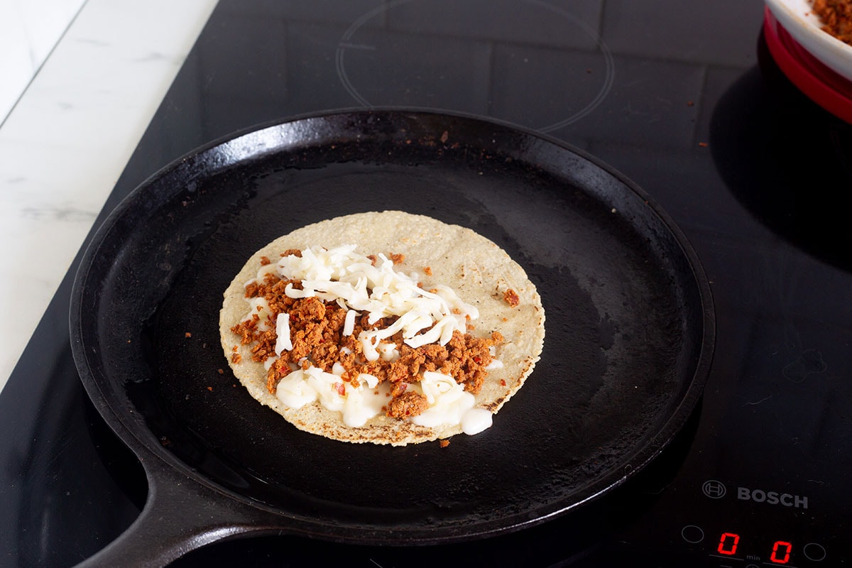 A corn tortilla topped with cheese and chorizo, on a griddle.