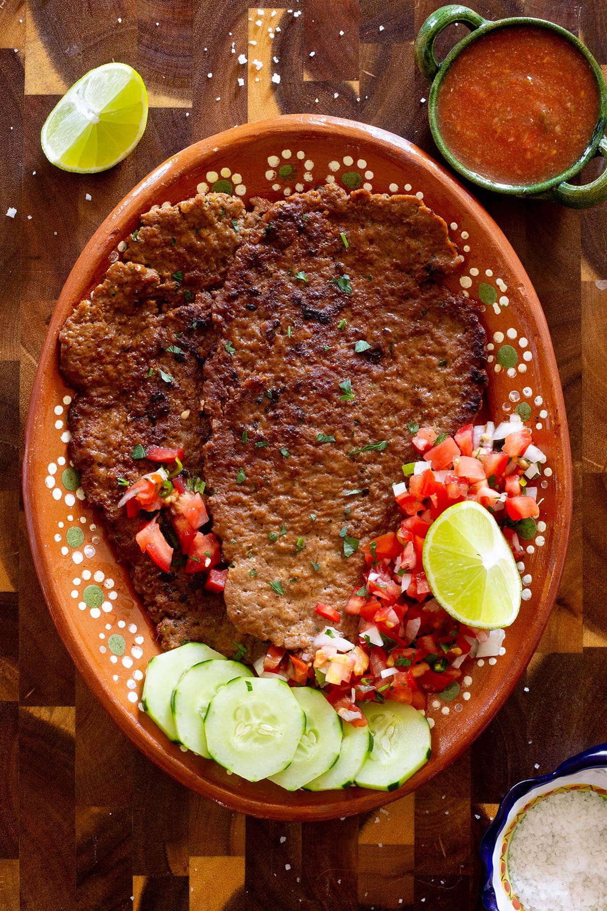 Beef Pacholas served with pico de gallo, salsa, cucumber slices, and lime wedges on a Mexican terracota platter.